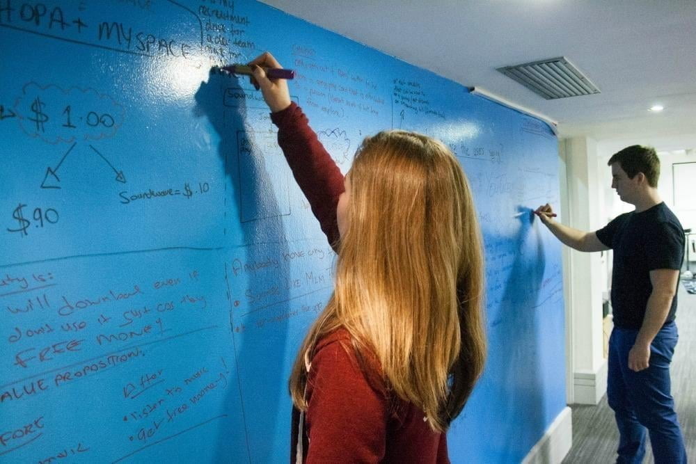 A woman and a man working together, writing on a blue whiteboard paint wall
