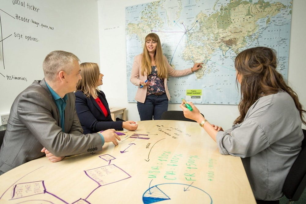 A group of people sitting at an office table which has been painted with clear whiteboard paint and they are writing on the table with whiteboard markers problem solving.